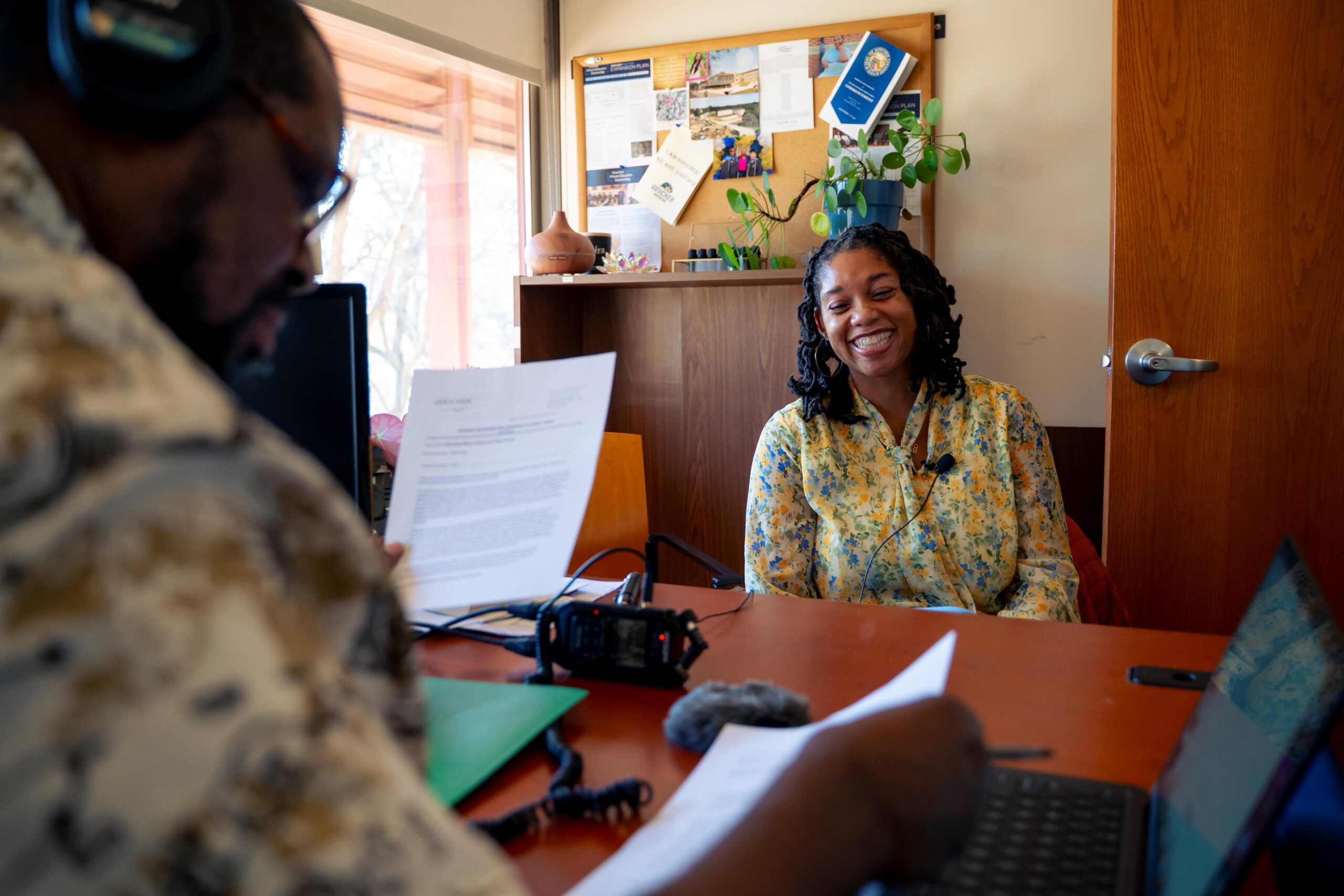 Woman smiles sitting at desk across from man with audio recorder and papers