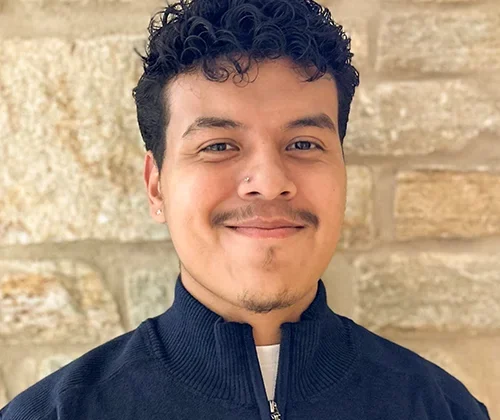 Nigel Cortez Cruz ’27 smiles in a navy blue quarter zip shirt in front of a stone wall on Goucher College's campus.