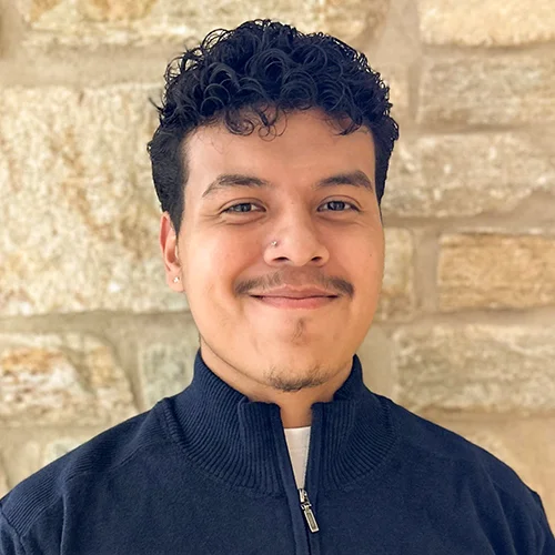 Nigel Cortez Cruz ’27 smiles in a navy blue quarter zip shirt in front of a stone wall on Goucher College's campus.