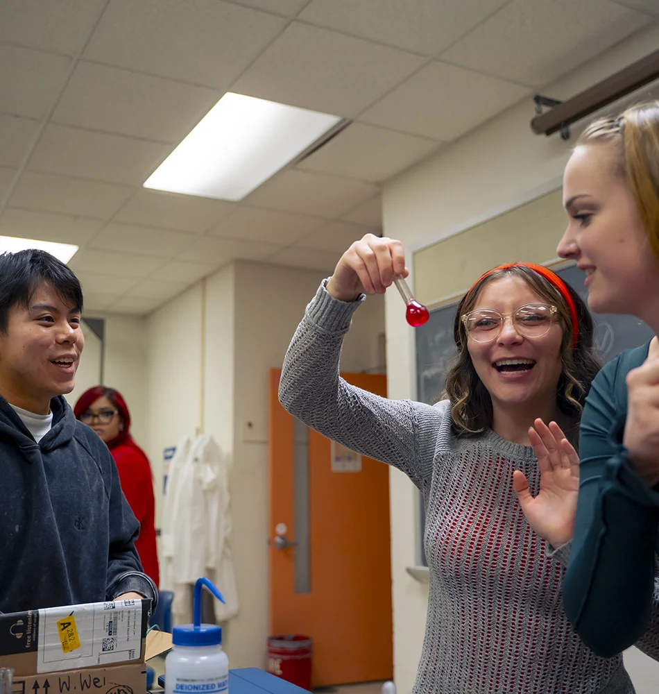 Students smile while one student holds a beaker in a hallway.