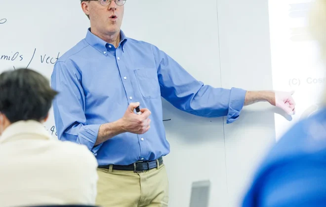 Prof. Mark Hiller stands at a white board teaching students in a classroom.