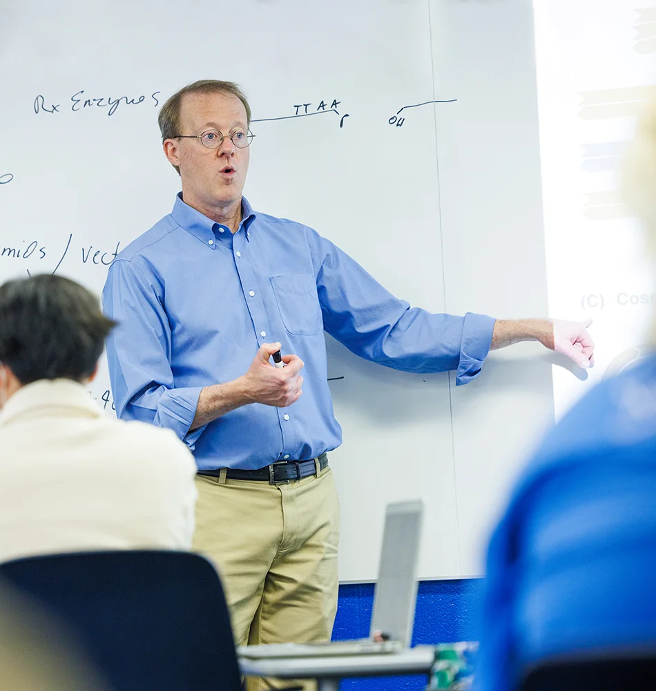 Prof. Mark Hiller stands at a white board teaching students in a classroom.